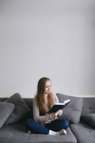 Smart Young Female Sitting On The Couch At Home Reading Book. Beautiful Woman With Long Hair In Mode