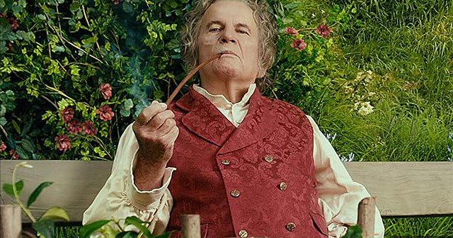 An older man with grey hair wears a red vest with a long-sleeved white shirt underneath. He sits in a garden while smoking out of a long pipe.