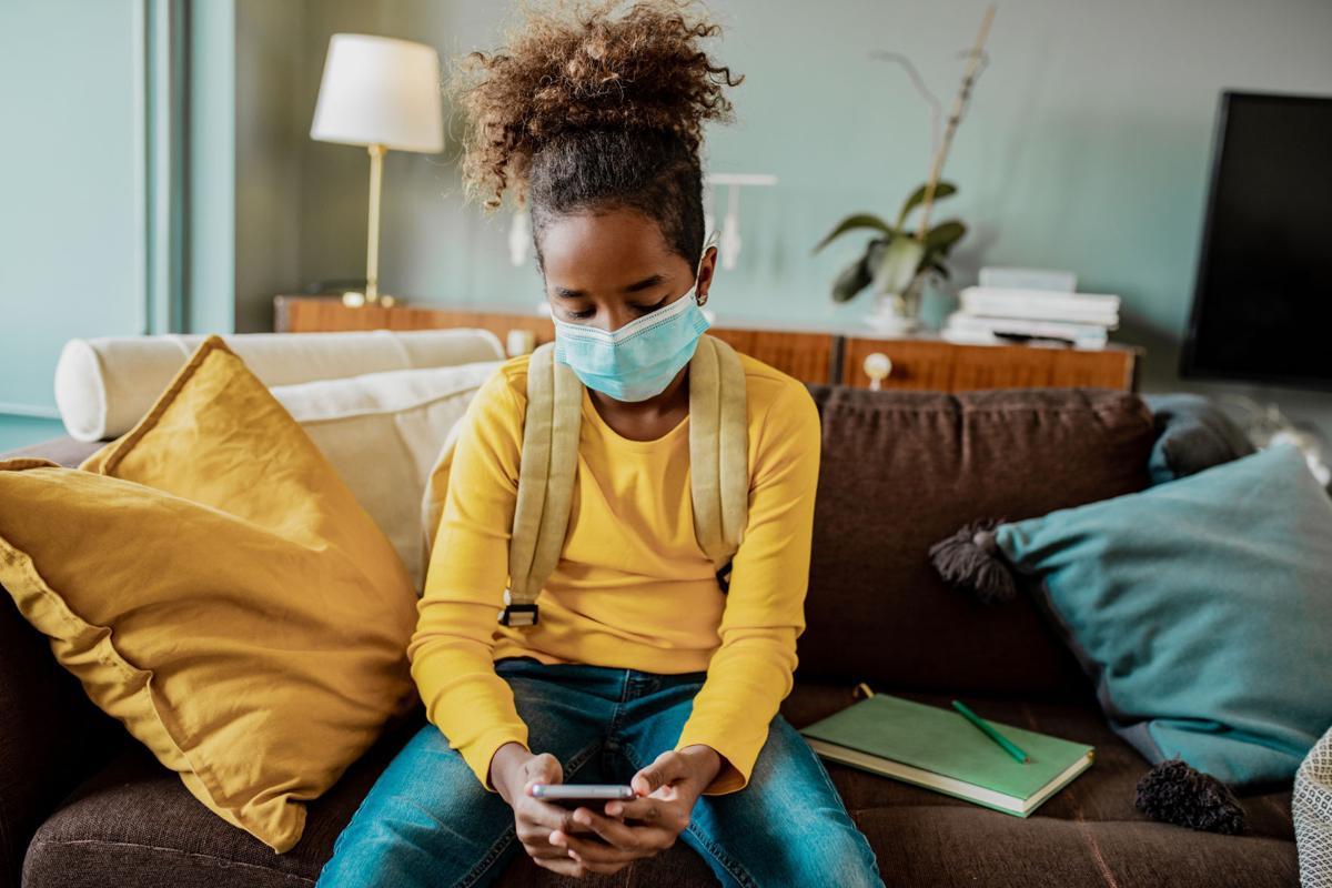 Sad African American girl with face mask at home using mobile phone