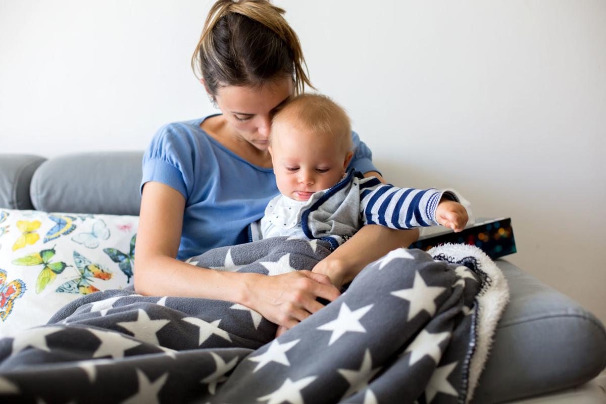 Young mother, holding her sick toddler baby boy in her arms, sitting on a couch in living room