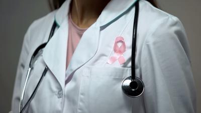 Lady doctor attaching pink ribbon to white medical suit, breast cancer awareness