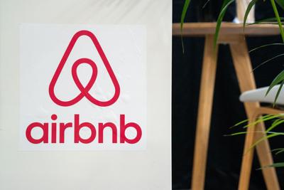 Red Airbnb Logo With Wordmark On A White Surface. In The Background, A Wooden Bar Stool And A Green Houseplant Are Visible.