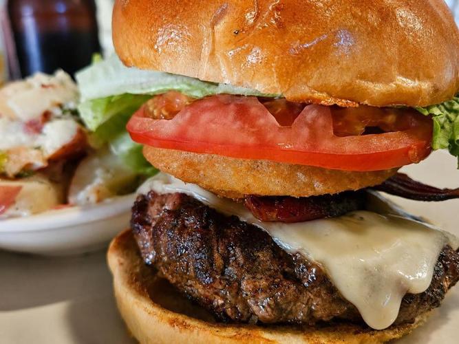 The Signature Steakhouse Burger at Alpine Steakhouse in Sarasota, Florida, is a half-pound patty stacked with house-made jowl bacon, a crispy onion ring, Swiss cheese, lettuce, tomato, and mayo. Photographed here with a side of German potato salad