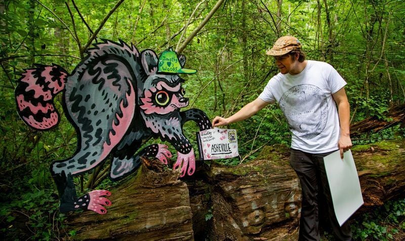 Artist Spencer Beals interacts with a painted depiction of Jimothy, the viral Seattle raccoon that gained internet fame in 2026, which he created for the Batland trail in Asheville on July 22, 2026.