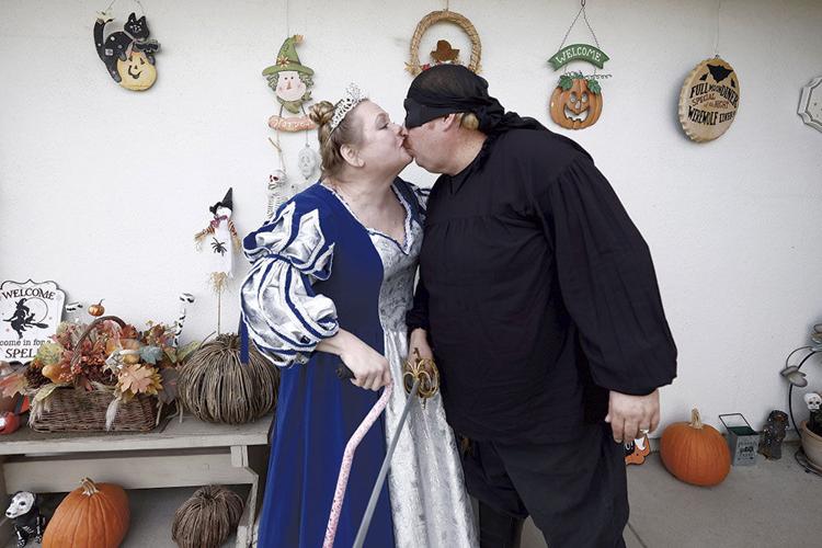 A white woman with blonde hair, wearing a silver crown and a blue-and-silver princess dress, holds a cane. She is kissing a white man on the mouth. He is dressed in all black and carries a fake sword.