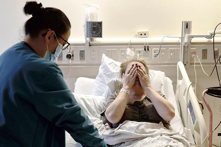 A woman sits in a hospital bed and holds her hands over her eyes, covering her face. A medical worker in scrubs stands next to her.
