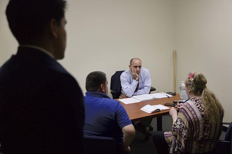 A bald white man wearing a button-down shirt and tie sits at a desk. He has a furrowed brow and looks across the desk at a woman sitting across from him. She has long blond hair with decorative flowers. She sits next to a man in a blue shirt.