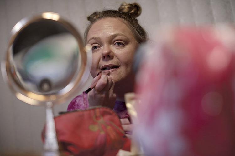 A woman’s face is shown close up as she uses her right hand to apply lipstick. She looks into a round mirror.