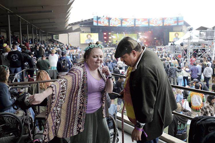 A woman and a man dance in front of a railing. Below them are hundreds of people standing or dancing, and a stage is viewable in the distance.