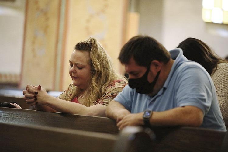 A white woman with blond hair sits on a wooden bench next to a white man with brown hair. They lean forward, as if in prayer.