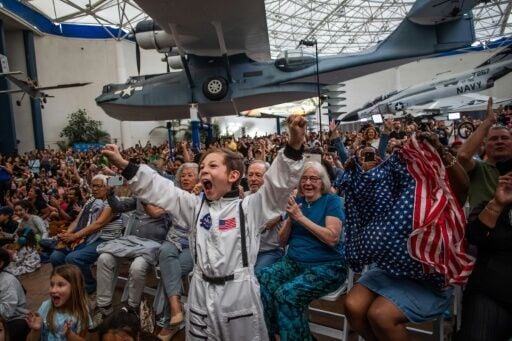 A young boy wearing an astronaut costume cheers next to a woman waving a flag as they watch a live broadcast of the return of the Artemis II crew members to Earth at the San Diego Air and Space Museum during a watch party