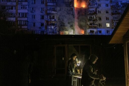 People walk past damaged shops in front of a residential building on fire following a Russian drone attack on the city of Vyshgorod, in the Kyiv region