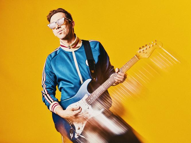 Cory Wong press photo artist in blue jacket and sunglasses plays a blue Fender Stratocaster against a yellow background