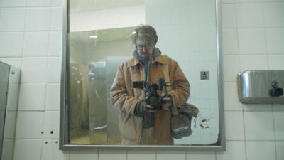 Still photo from 'The History of Concrete,' in which director-host John Wilson, dressed in construction safety gear, aims his camera into the mirror in a public restroom coated in concrete dust.