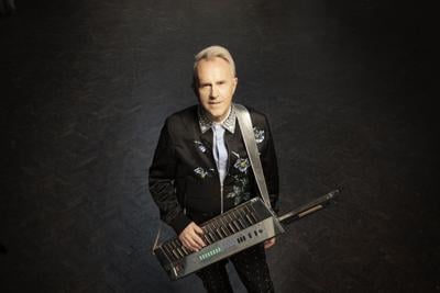 Howard Jones press pic artist wears a dark color Western shirt holds a keytar and smiles at the camera