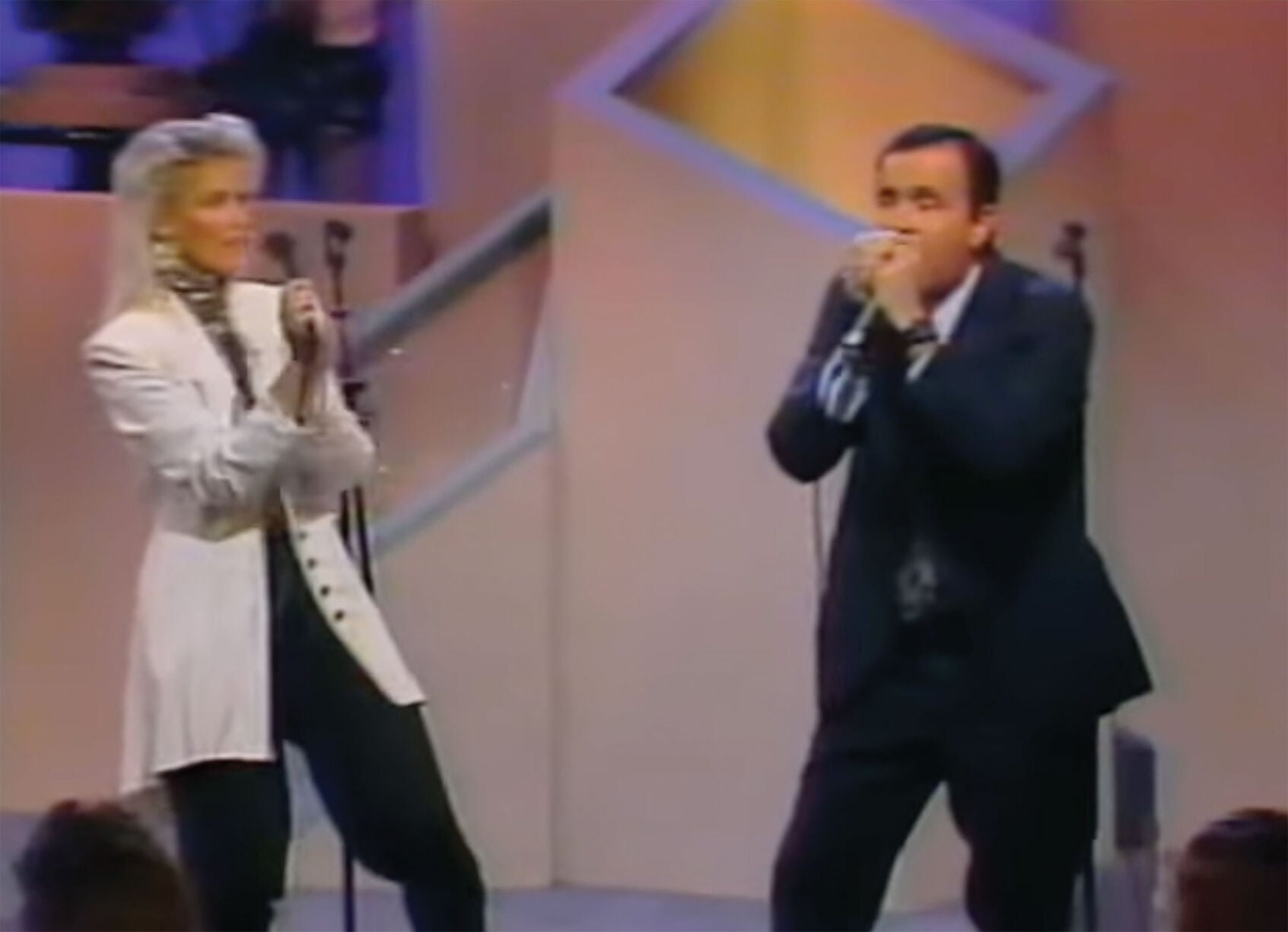 A woman in a white jacket claps as a man in a dark blue suit plays the harmonica into a microphone. Both are on stage.