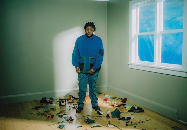 Color photo of Joy Oladokun standing near an uncovered window in a corner of a room amid a pile of flowers spilled from a broken vase and pieces of a broken LP record