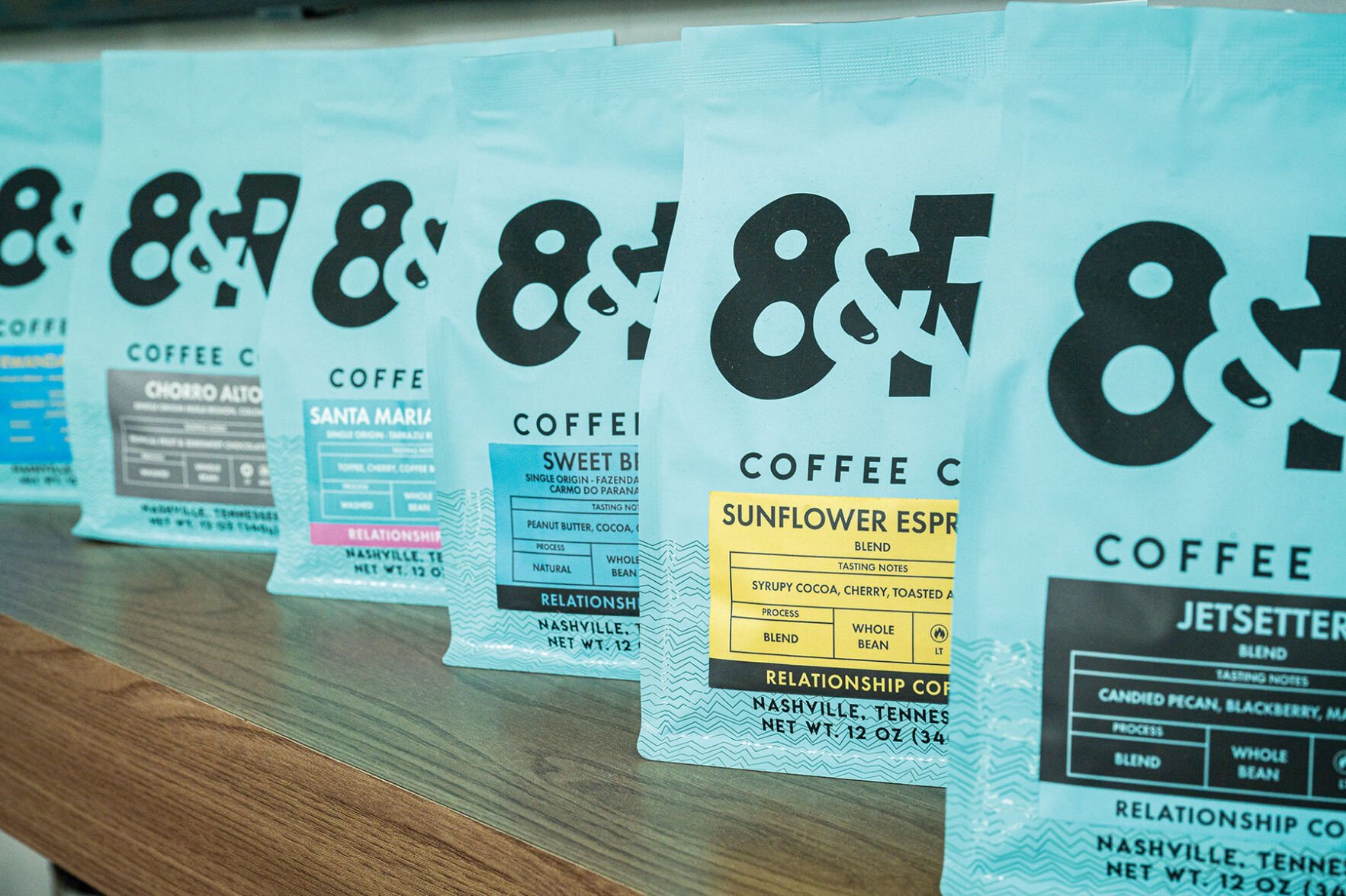 A row of light blue 8th & Roast Coffee bags with black, yellow and blue stickers indicating the different types of roasts and blends in each one.
