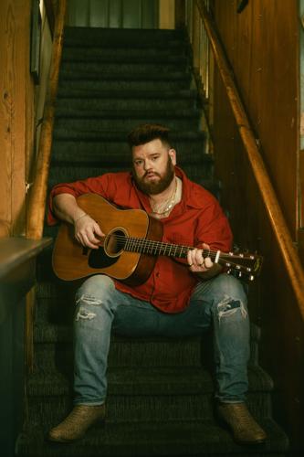 Color portrait of the artist in a red button-up shirt, jeans and cowboy boots playing an acoustic guitar on the stairs inside Margot Cafe & Bar