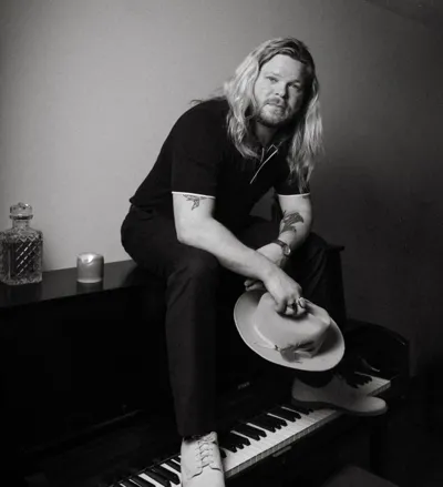 Black and white press photo of John Countryman, the Nashville Scene’s 2026 Best Instrumentalist, sitting atop a piano and looking at the camera