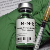 Green medical gloved hand holding a MMR vaccine vial