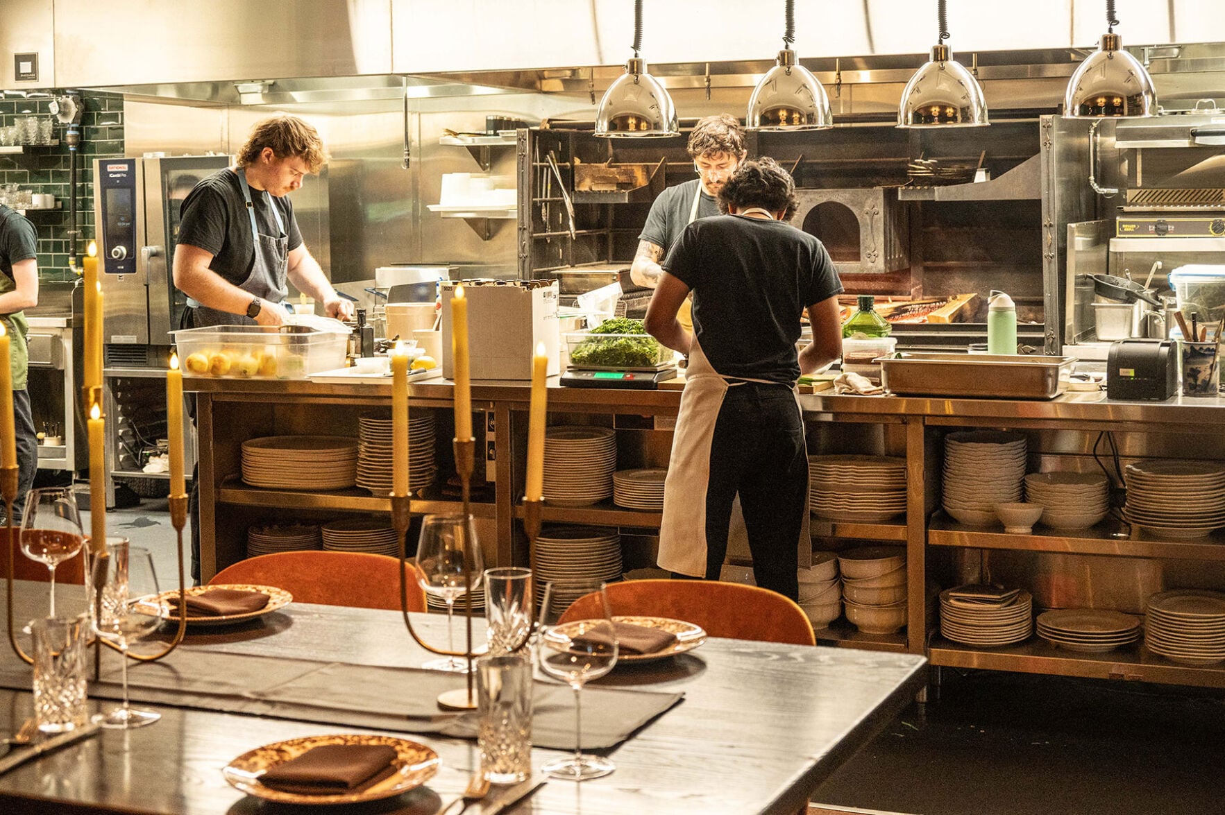 Three restaurant works hunch over a large counter, plates stacked beneath in shelves, with a dining table in the foreground.