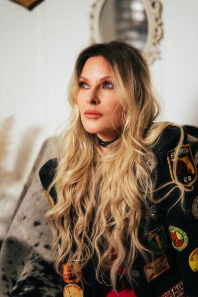 Color press photo of Elizabeth Cook, wearing a jacket covered in travel patches and looking above and to the right of the camera