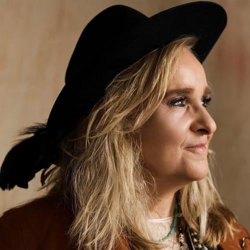 Color press photo featuring a closeup headshot of Melissa Etheridge, wearing a broad-brimmed hat with a feather in the back of it and looking off to the right.