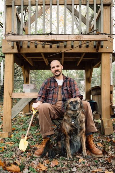 Color press photo of the artist and his dog sitting outdoors under a deck, surrounded by autumn leaves