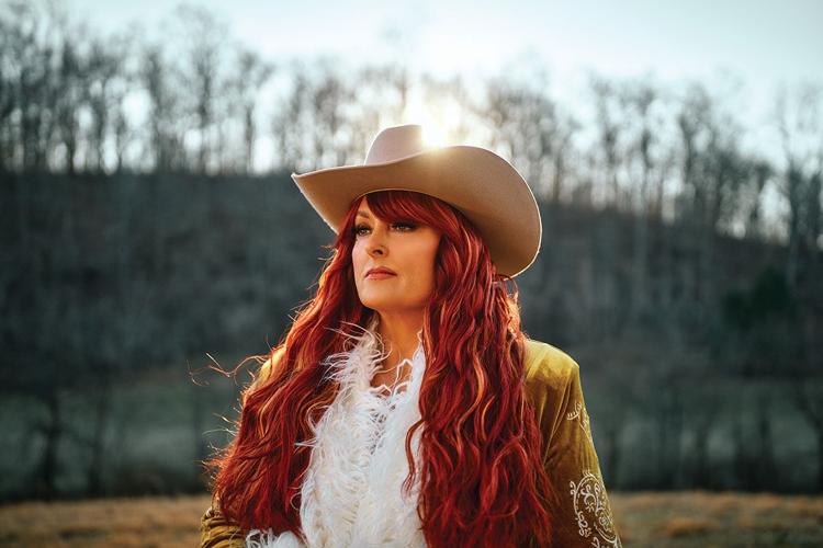 Color press photo of Wynonna Judd, wearing a cowboy hat and standing outside with the sun setting through the trees behind her, looking off to her right.