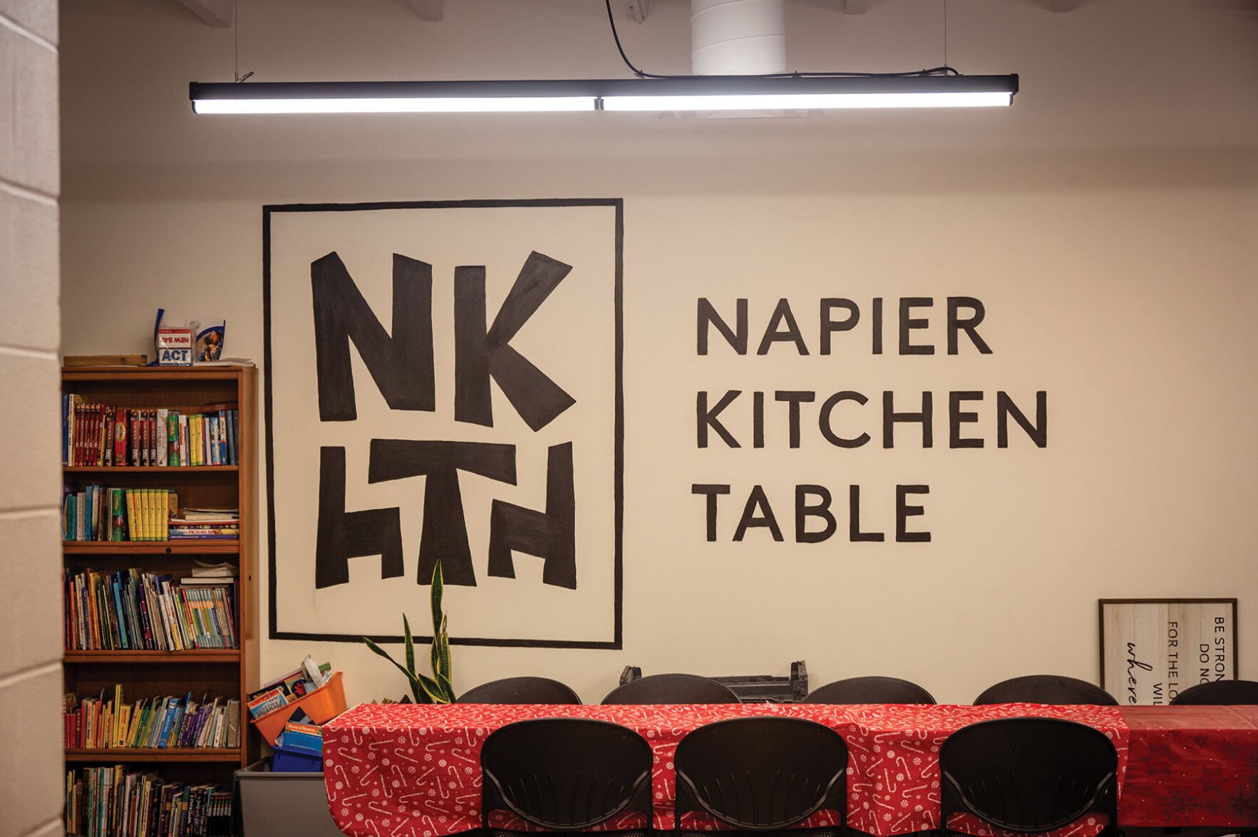A mural on a wall reads "Napier Kitchen Table"; beneath it stands a table covered in cloth with chairs arranged around it.