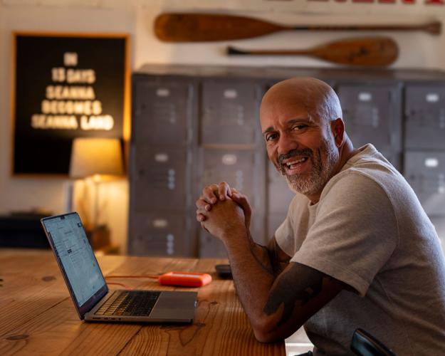A man in a white t-shirt sits in front of an open laptop, elbows on the table and hands clapsed, with a set of lockers behind him