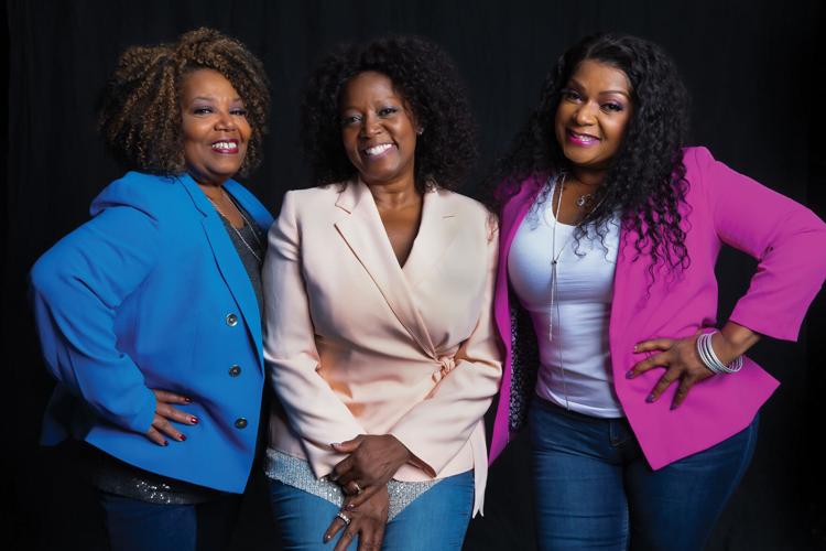 Color photograph of Freda, Ann and Regina McCrary, wearing blazers and jeans and standing against a charcoal gray background