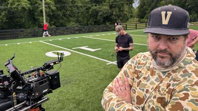 A man in a Vanderbilt baseball cap and camo shirt stands next to a camera on a football field, members of the film crew are scattered in the background