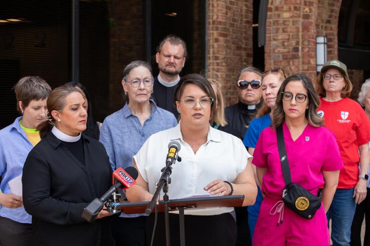 A woman in a white shirt with glasses speaks at a lectern, a crowd behind her, including a nurse and a reverand.