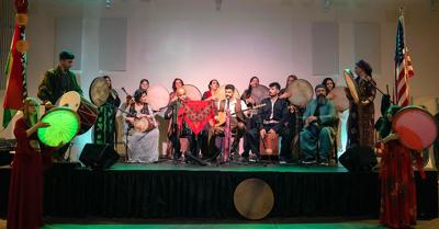 Color photo of the Rhythm Group ensemble from the Kurdish Culture and Music Association performing onstage, flanked by Kurdish and United States flags.