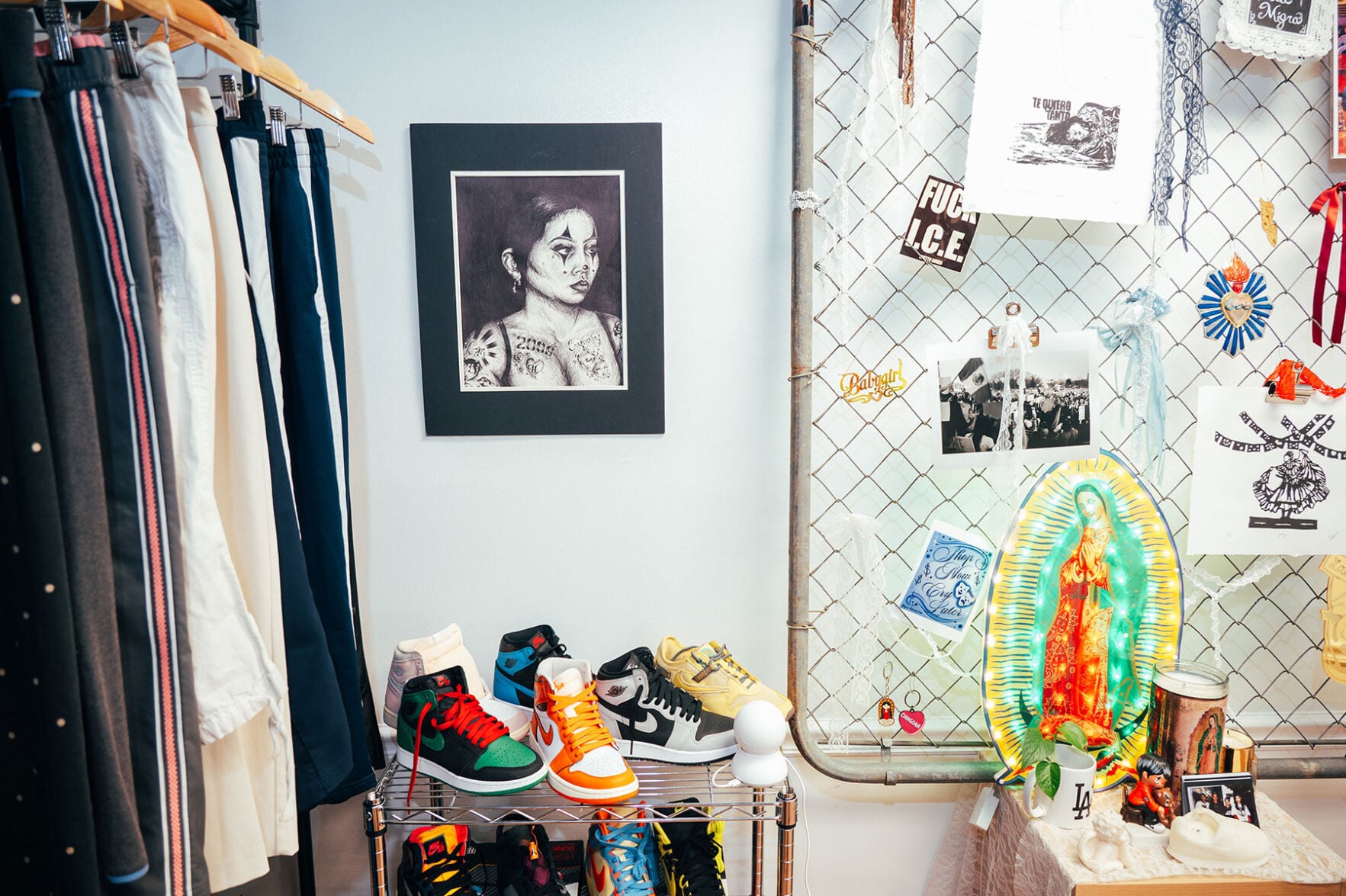 A collection of jackets and sneakers next to a black and white photo of a tattooes woman and a poster board covered in stickers and drawings, including a colorful depiction of the Virgin of Guadalupe
