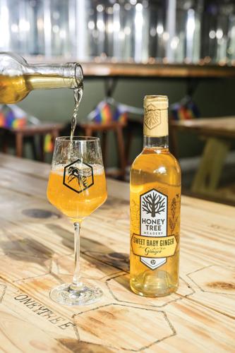 A bottle of Sweet Baby Ginger from Honeytree Meadery placed next to a glass that is being filled by another bottle poured from above it.