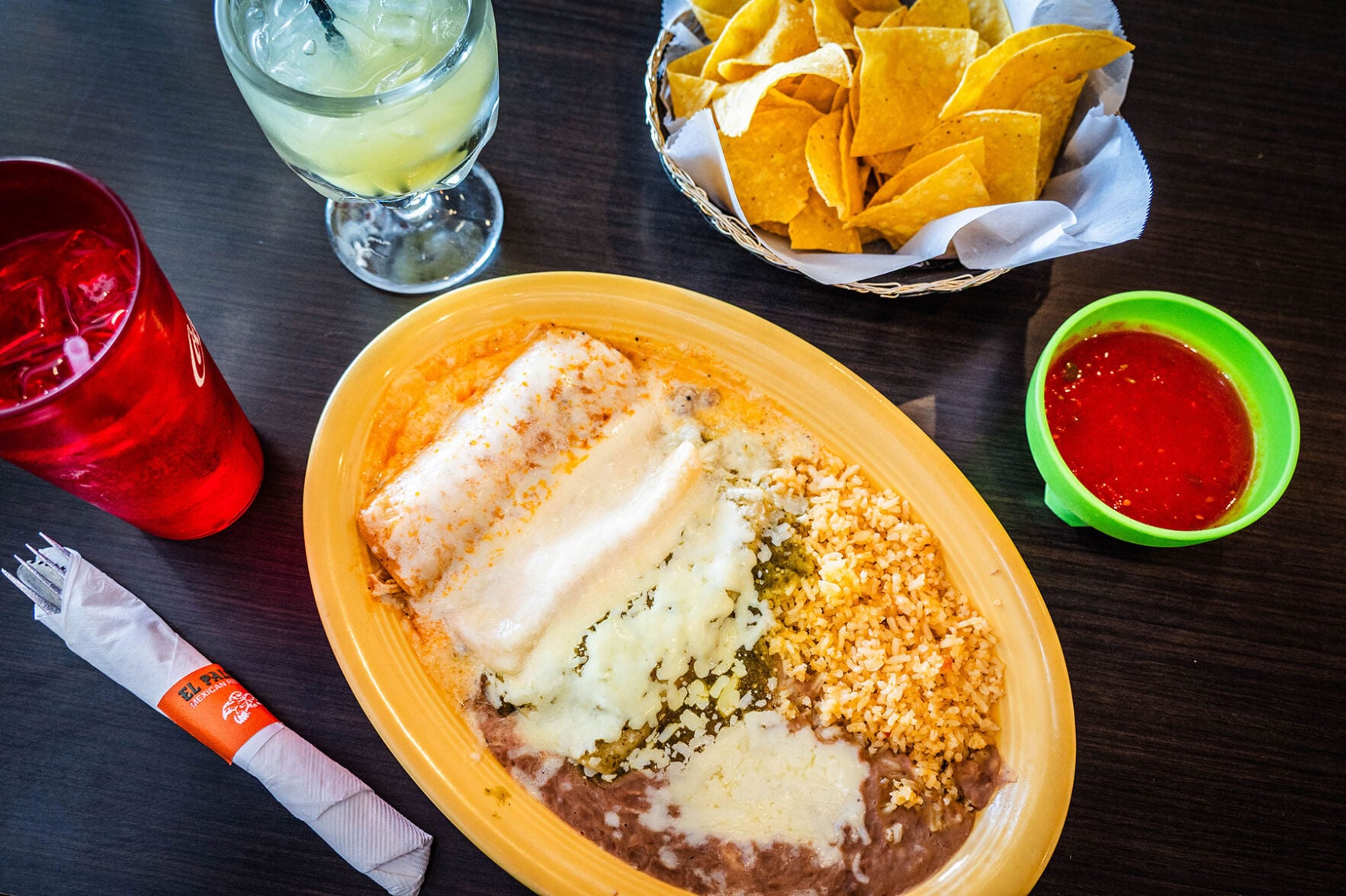 Cheese-smothered enchiladas next to rice and beans on a yellow plate, with a bowl of salsa and basket of chips nearby, along with a margarita