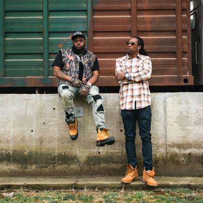 Color press photo of producer Bandplay and MC Starlito hanging out by a loading dock on a farm, sporting flannel, camo and unlaced work boots.