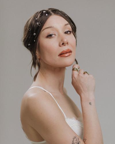 Artist poses against a neutral background, wearing a white gown and iridescent decorations woven into her hair.