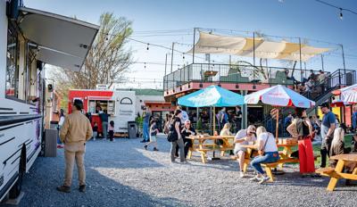 At the far right view, a man orders from a food truck, with another visible in the distance advertising empanadas. A few picnic benches with ubrellas are to the left, in front of a two floor structure that has a deck on top.