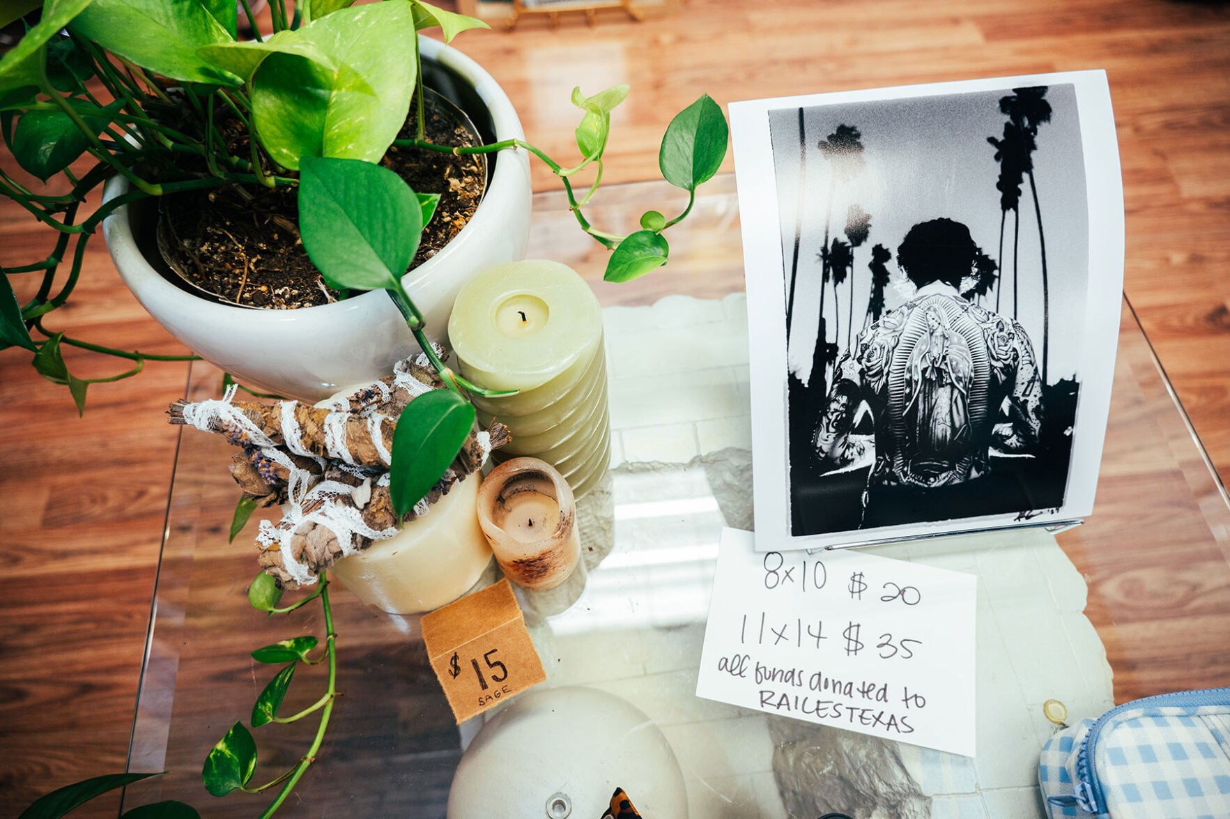 A glass table holds a potted plant, a few candles priced at $15 and a black and white photo of a man with an intricate back tattoo. A sign next to the photo lists prices: 8 by 10 for $20 and 11 by 14 for $35, with all funds donated to Raices Texas.