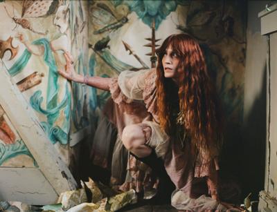 Color press photo of Florence Welch, in makeup with a sunken eye effect, emerging from a pile of rags in the corner of a dimly lit room with a mural of demons on the walls