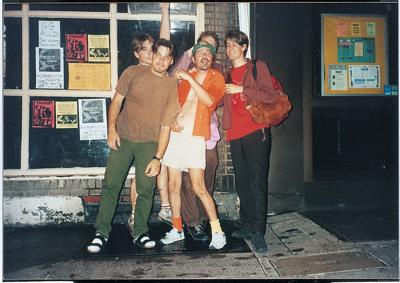 1990s color press photo of the rock band Pavement, standing together outside a record store or music venue with posters in the window