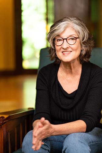 Color author photo of Barbara Kingsolver, dressed in a black top and jeans, sitting on a chair in a room with a wood floor with an open door behind her, pressing her hands together in a neutral stance and looking into the camera.