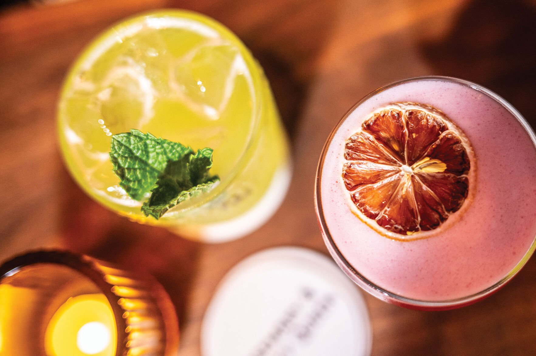 Photo taken from above of a yellow drink garnished with mint and a pink drink garnished with dried orange