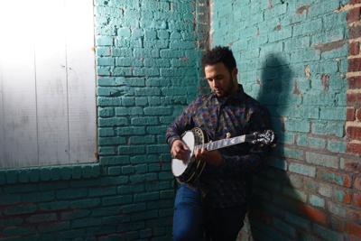 Standing against a brick wall painted green, the artist poses in a flannel shirt playing a banjo.