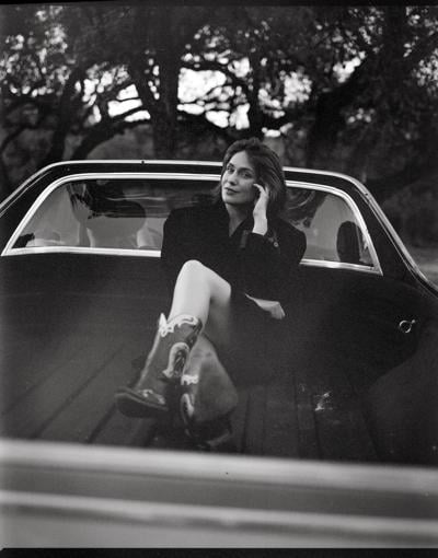 Black and white photo of musician, author and actor Lola Kirke, wearing a dark dress and cowboy boots and seated in the bed of a vintage truck with her back against the cab.