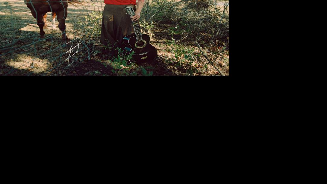 Holding a guitar and wearing dark pants and a bright red T-shirt, artist reaches across a fence to scratch a horse's chin